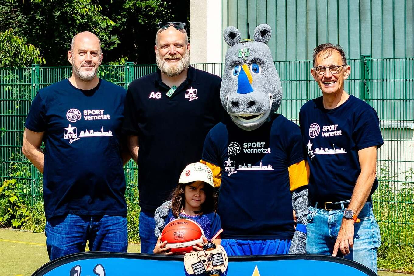 Vier Personen und ein Maskottchen stehen vor einem Banner der TVE Barop, umgeben von grüner Natur.