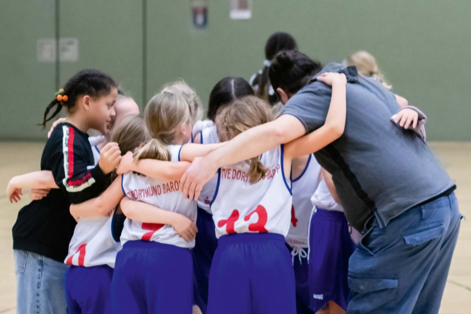 Mädchen-Basketballmannschaft umarmt sich vor einem Spiel, Trainer steht in der Mitte, alle tragen einheitliche Trikots.