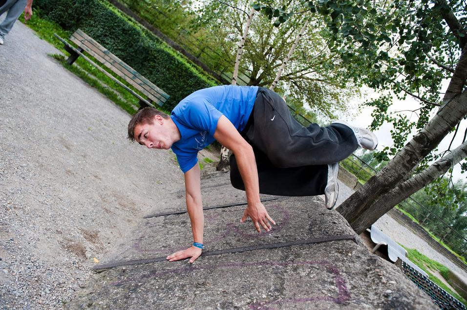 Junger Mann führt eine Parkour-Bewegung auf einer Mauer in einem Park aus, mit Bäumen und einer Bank im Hintergrund.
